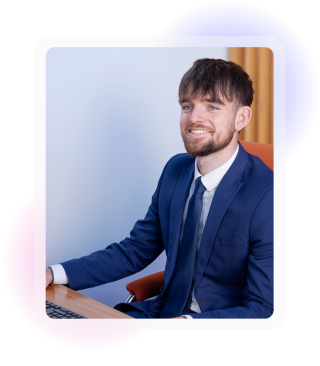 Young male in office attire, sat at a desk with laptop and smiling. 