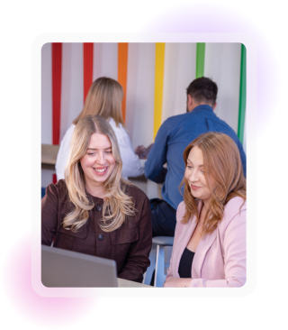 Two female employees in office attire sat at a desk looking at and discussing something on the laptop