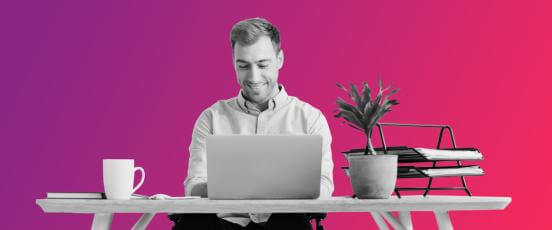 A man sat at a desk with a laptop, book, coffee cup, plant and file system on top. The man is working happliy remotely after using People First's Workforce management solution software.