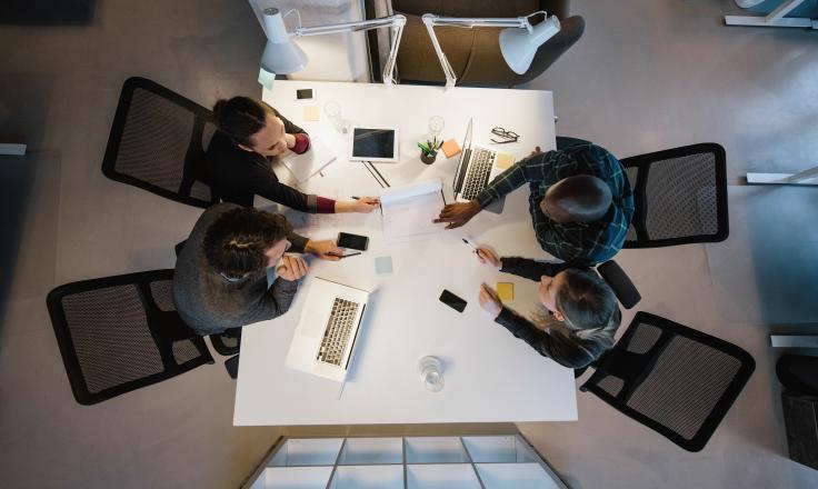 4 people sat around an office desk having a meeting, representing business as usual