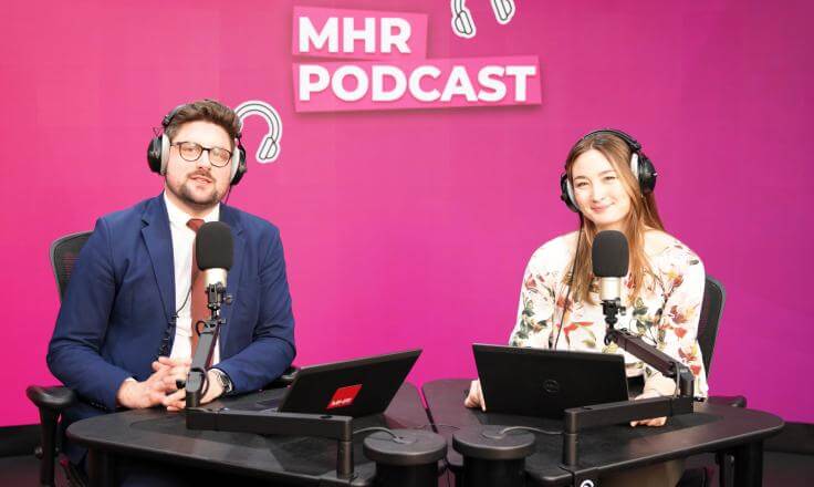 Alice and Andy sat with headphones on in front of a mic and laptop, with MHR podcast written behind them.