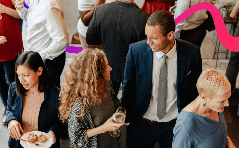 A group of professionally dressed people networking at a business event, with attendees chatting while holding drinks and light refreshments