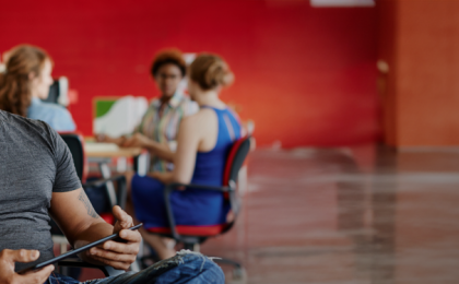 A man sat smiling with a tablet on his lap, with a group of people behind him at the table. Happy about integrating Learning Management System onto their People First platform.