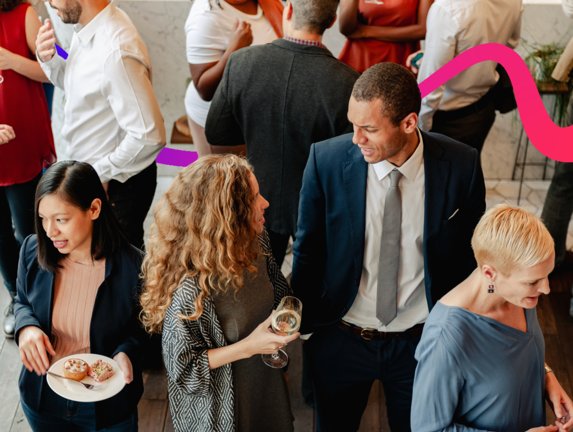 A group of professionally dressed people networking at a business event, with attendees chatting while holding drinks and light refreshments
