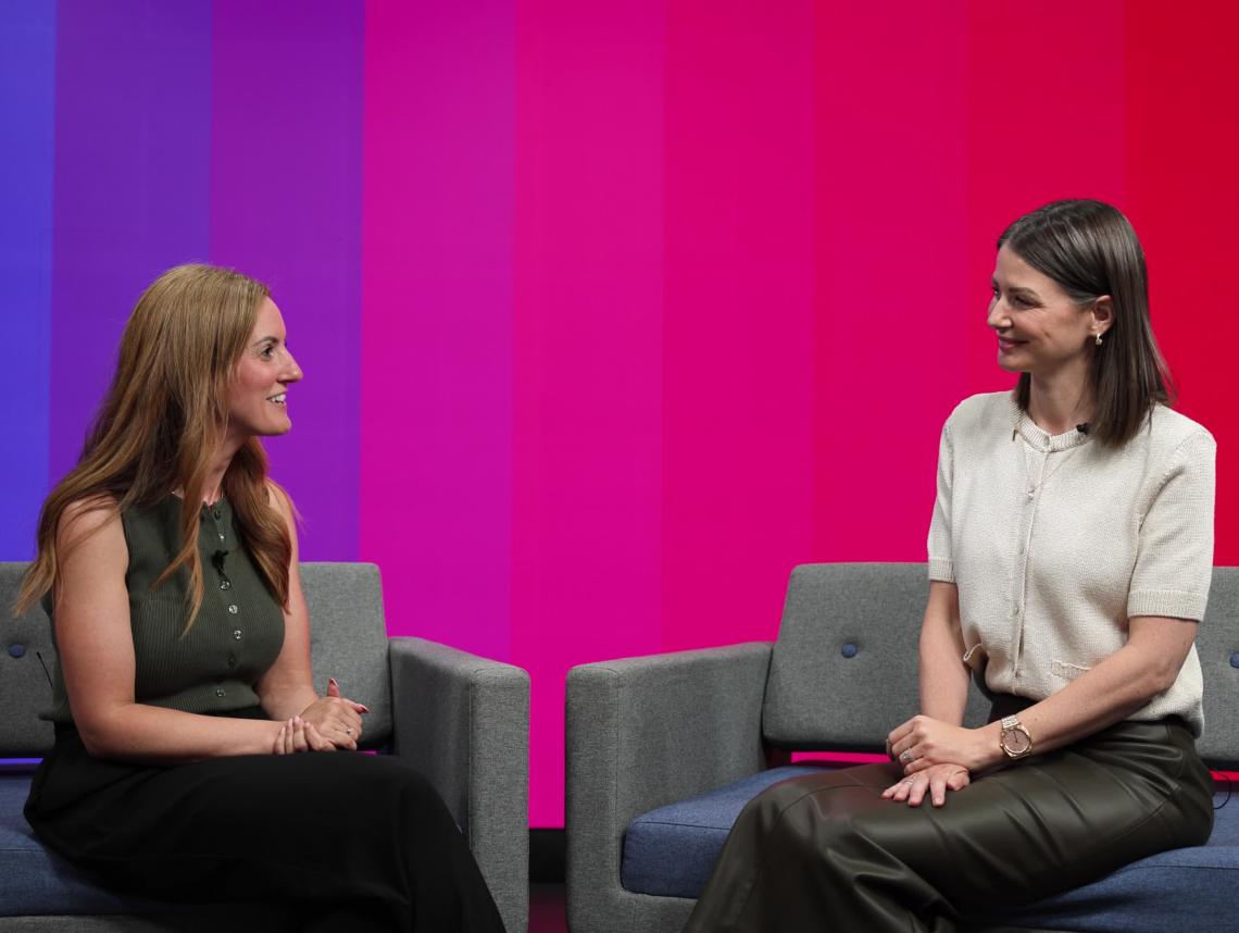 Two women sit on sofas in front of a colourful background.