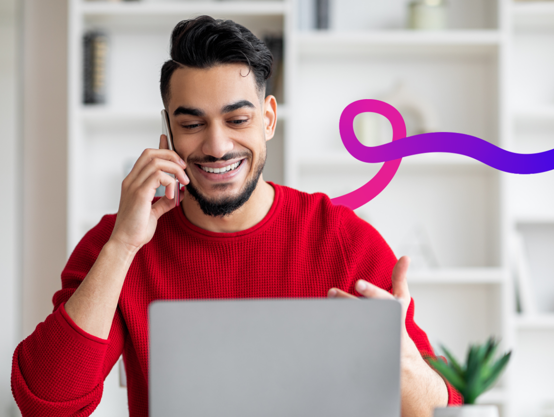 A man smiling with his phone to his ear, looking at a laptop