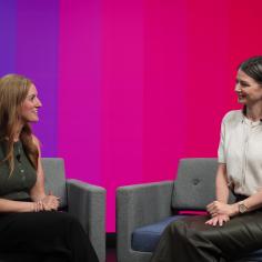 Two women sit on sofas in front of a colourful background.