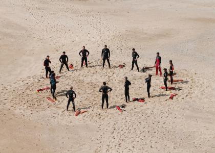 14 lifeguards wearing wet suits, stand in a circle, on a sandy beach
