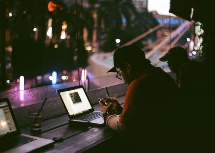 Two people sit at a long wooden table with laptops, facing a window with neon signs outside 