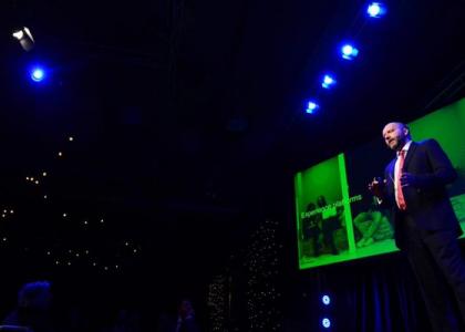 A man on a stage giving a speech with a presentation on a screen behind.