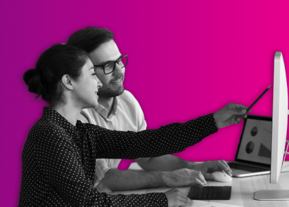 A man with glasses using a computer mouse, and a lady pointing a pen at a desktop, sat looking at analytical data.