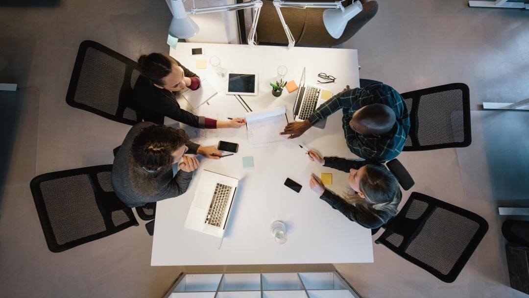 4 people sat around an office desk having a meeting, representing business as usual