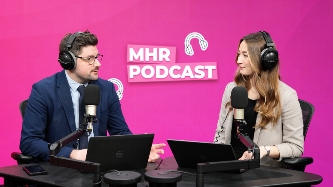 Andy and Alice sat with headphones on infront of a podcast mic, looking at each other with laptops in front of them.