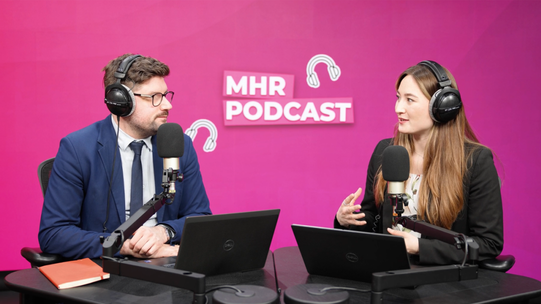 Alice and Andy sat with headphones on infront of a mic and laptop, with MHR podcast written behind them.