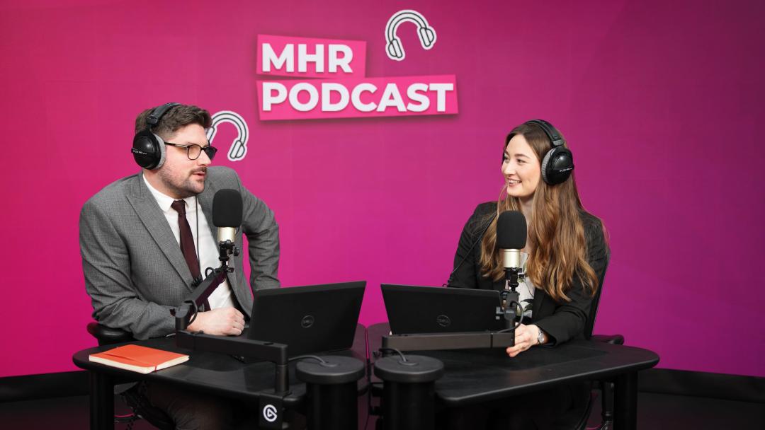 Alice and Andy sat with headphones on infront of a mic and laptop, with MHR podcast written behind them.