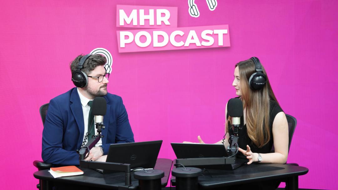 Alice and Andy sat with headphones on infront of a mic and laptop, with MHR podcast written behind them.