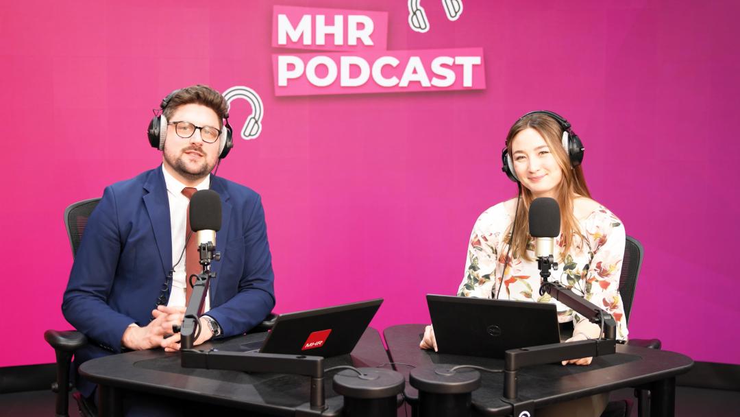 Alice and Andy sat with headphones on in front of a mic and laptop, with MHR podcast written behind them.