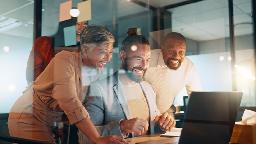 Three people sat around a laptop in an office, smiling.
