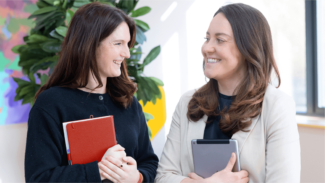 Two colleagues holding notepads smiling at each other in an office setting
