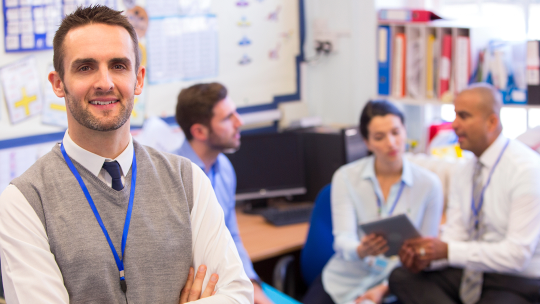 School staff member standing in a classroom while colleagues collaborate in the background.