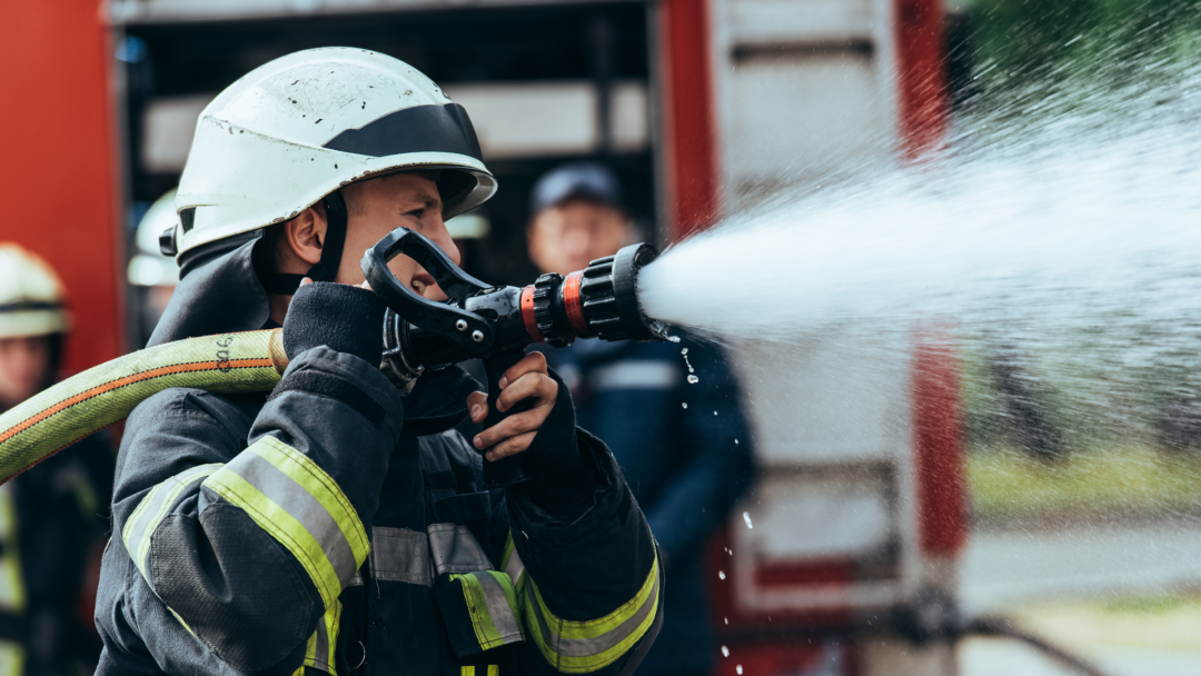 Firefighter in protective gear directing a powerful stream of water form a hose beside a fire engine,