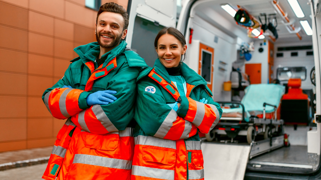 Two smiling paramedics in high-visibility uniforms standing in front of an open ambulance.