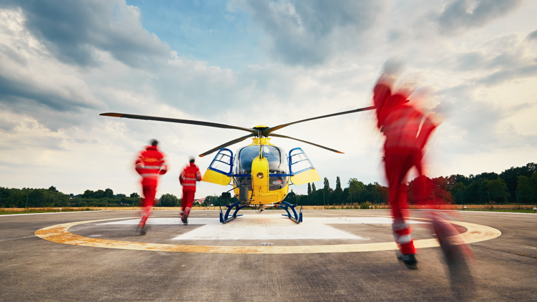 Yellow rescue helicopter on a helipad, with three emergency responders in red uniforms running toward it.