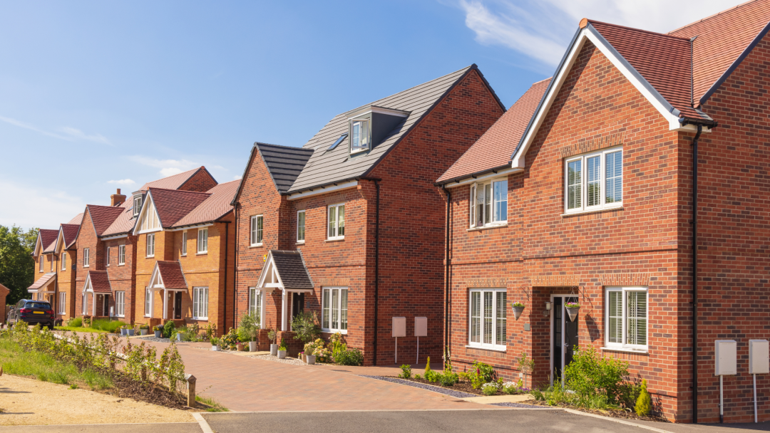 Row of newly built red-brick houses with landscaped front gardens under a blue sky.