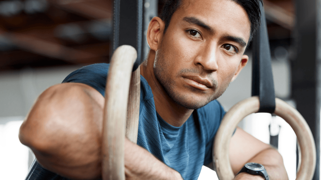 Close-up of a man in a blue shirt exercising on gymnastic rings, looking toward the camera.