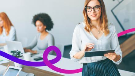 Woman sat on the edge of a desk holding an iPad. Two other women sat in the background.