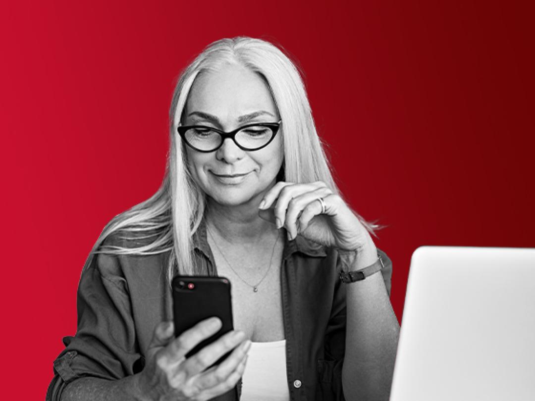 A lady with glasses, using a mobile, sat at her laptop. Happy after using iTrent for the MCR pensions.
