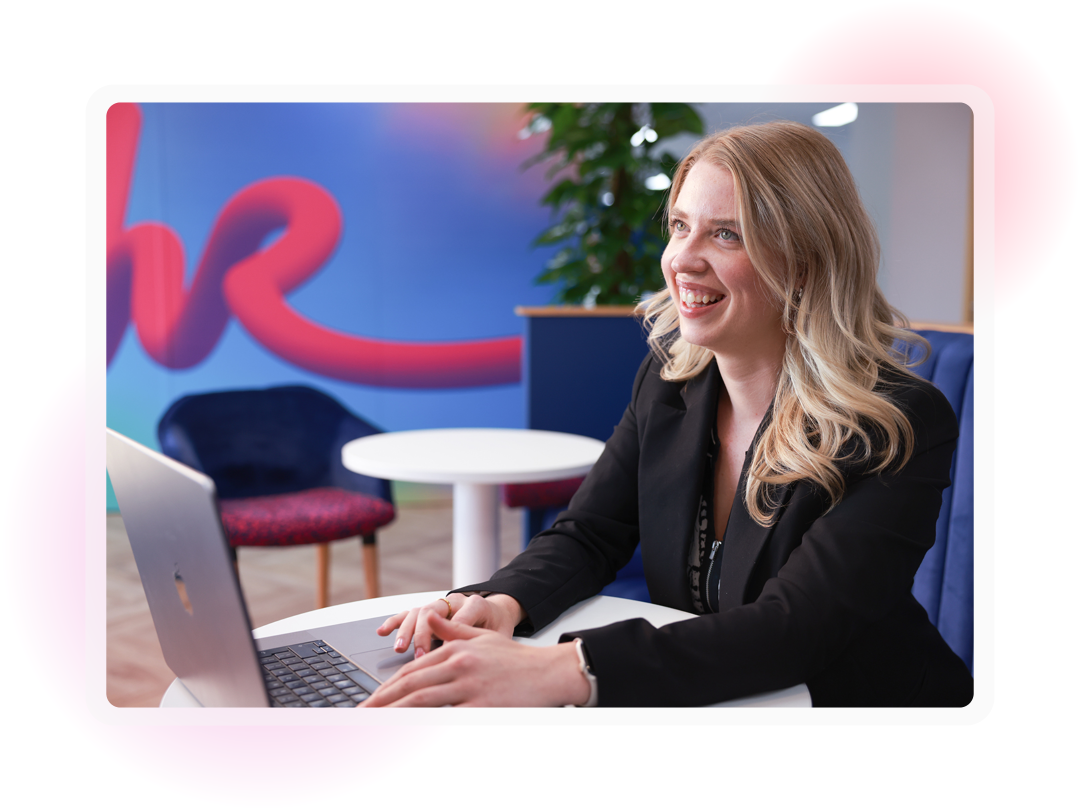 A young female sat in an office environment with a laptop on a table infront of her